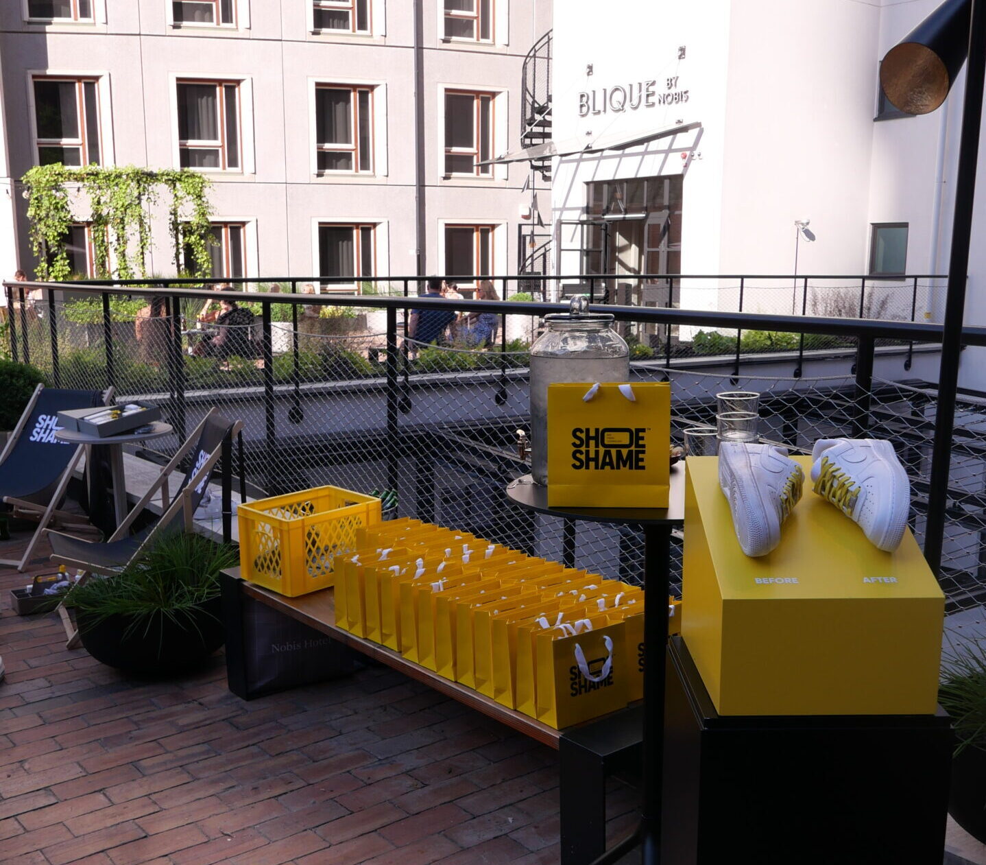 Innergård med växtlighet, gula Shoe Shame-påsar uppradade på en bänk med skor och produkter intill.